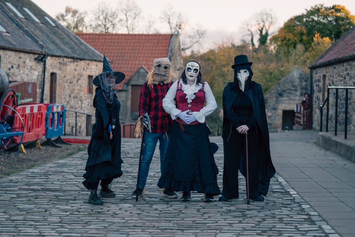 Four people stand in the main street at Almond Valley, they are all wearing Halloween style costumes.