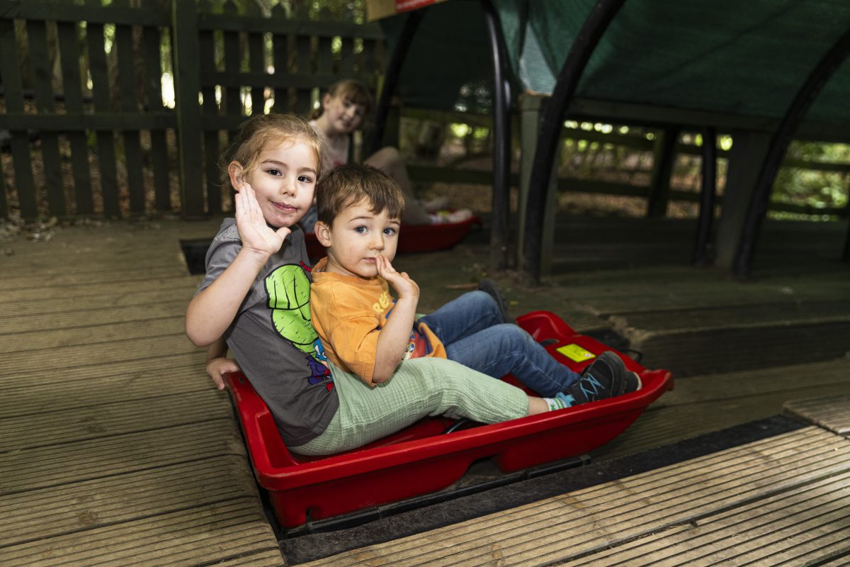 Two young children sit on sleds at the top of the slide. They are waving.