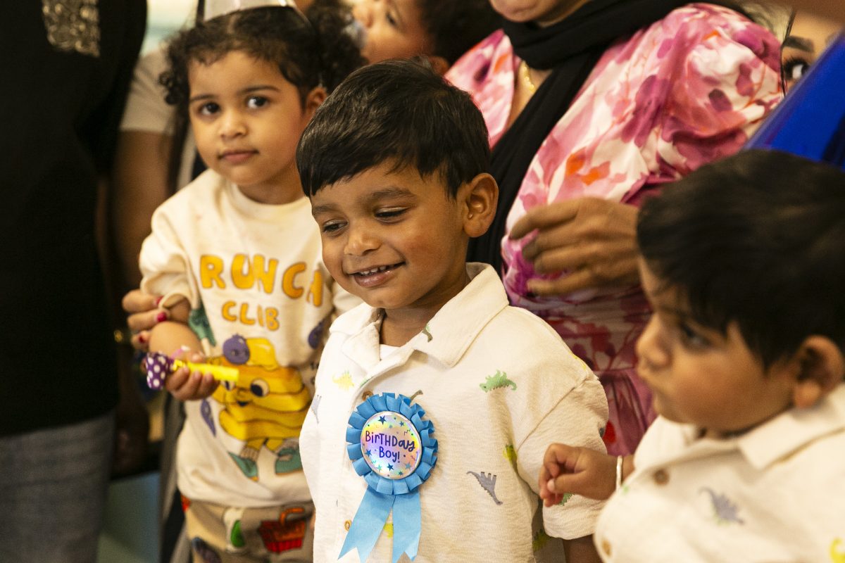 Three young children gather at a party. The one in the middle wears a rosette that says birthday boy.